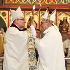 Dwóch biskupów w białych szatach i mitrach podczas ingresu do katedry. Jeden przekazuje drugiemu pastorał.