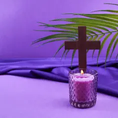 Drewniany krzyż, płonąca świeca i gałązka palmy na fioletowym tle. Symbolika postu i refleksji.
