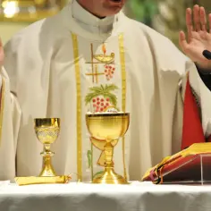 Kapłan w uroczystej szacie z haftem winogron i hostiami, podnosi ręce. Obok kielichy i księga, na której widnieje napis "Credo".