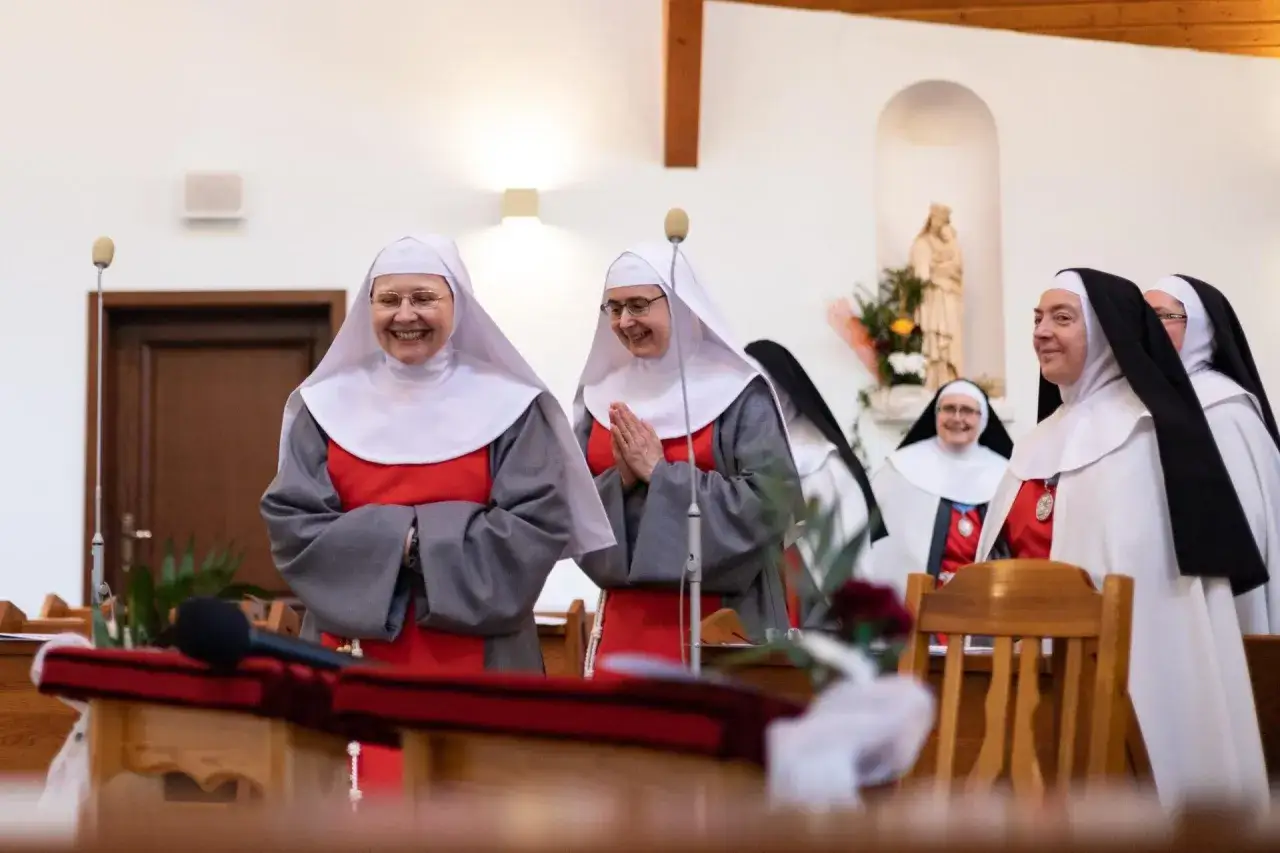 Uśmiechnięte zakonnice w białych habitach, jedna z nich z dłońmi złożonymi do modlitwy, w kościele.